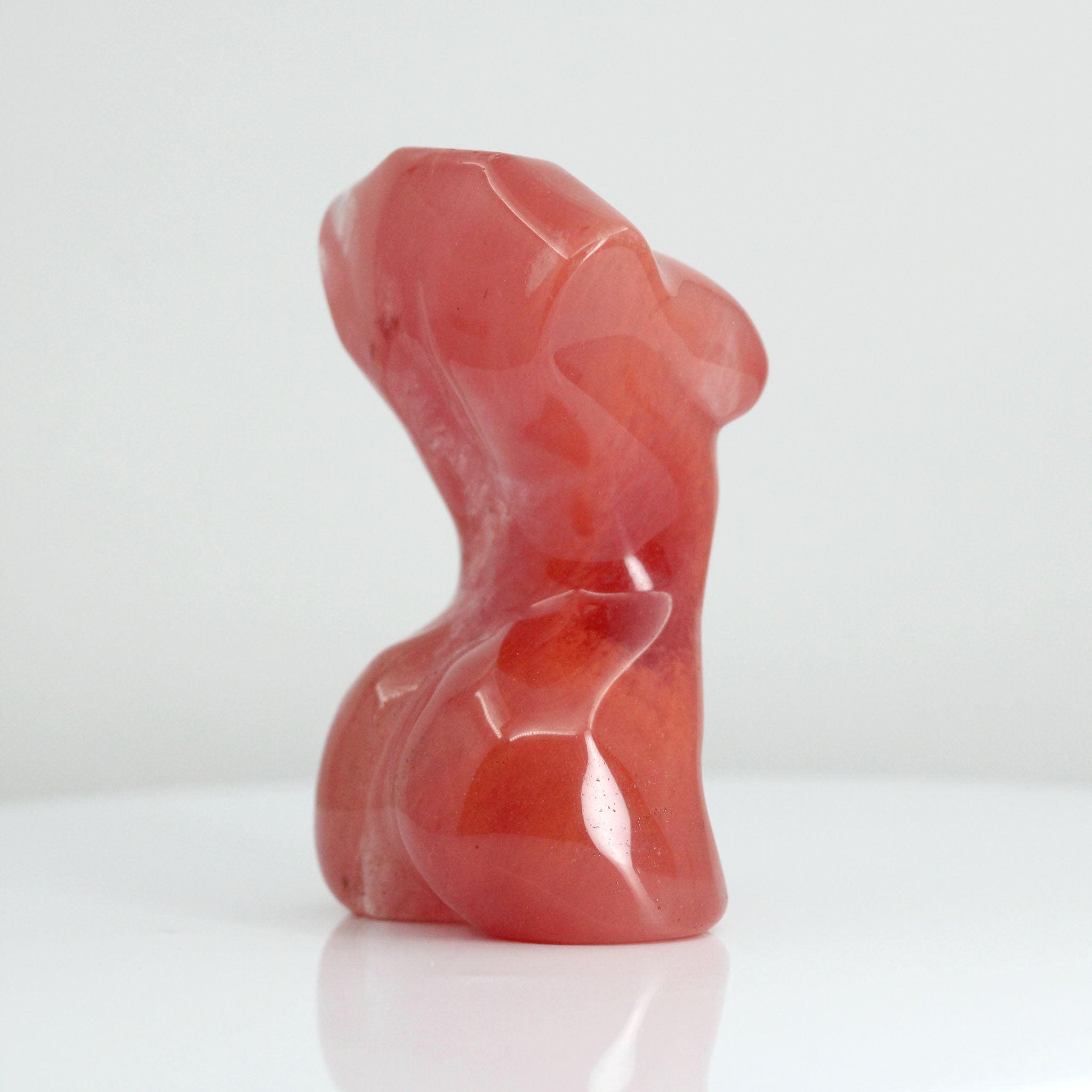 Side view of a sculpture in the shape of female body from neck down to upper thigh, with no arms, made from cherry quartz on a white background