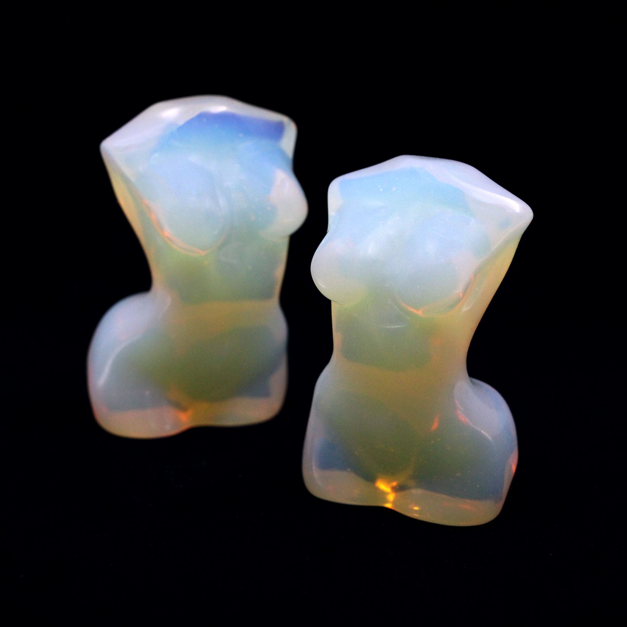 Two sculptures in the shape of female bodies from neck down to upper thigh, with no arms, made from opalite, a man-made opalescent glass that is milky white with subtle blue and iridescent qualities, on a black background
